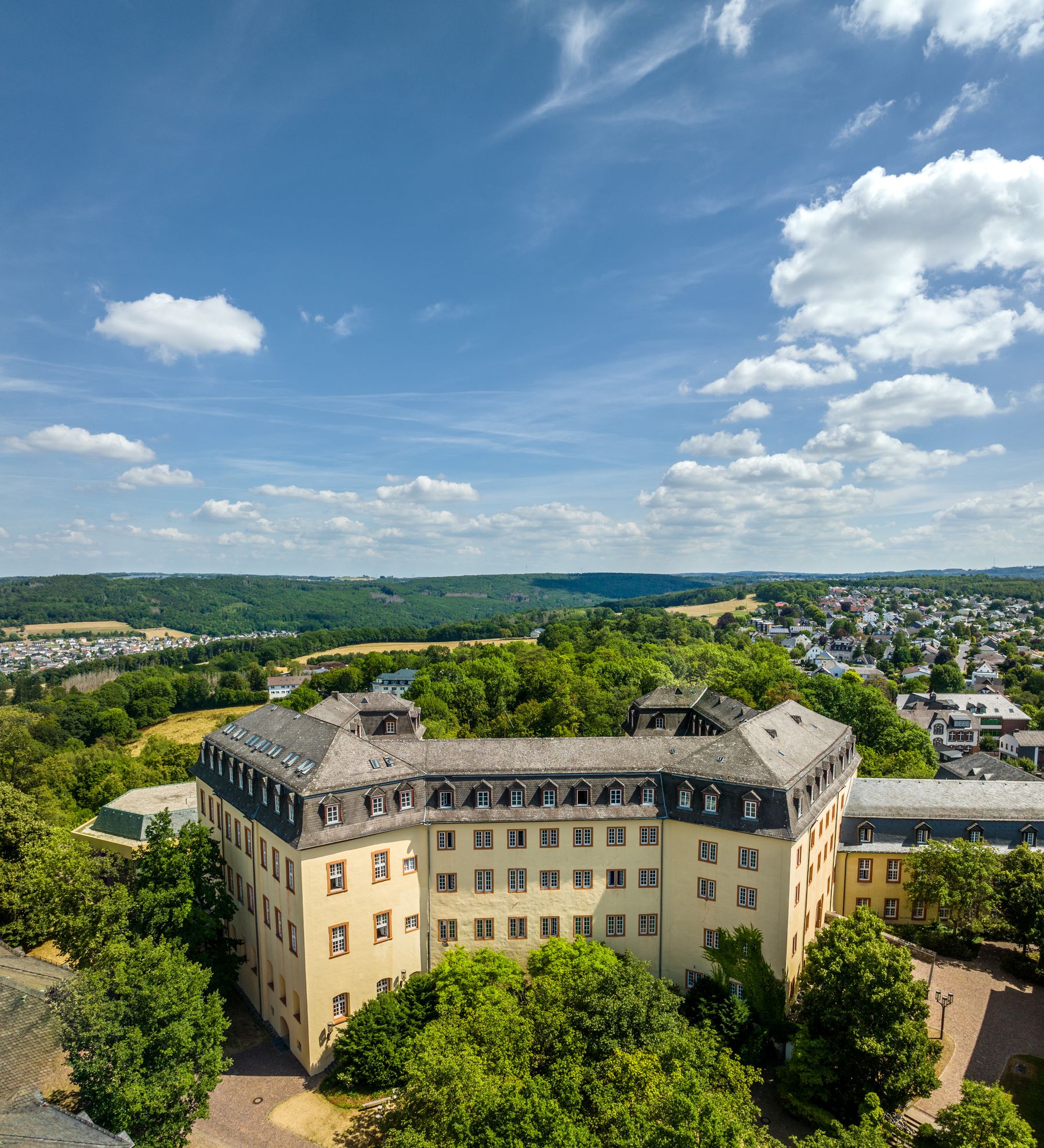 Luftaufnahme eines großen, historischen, blassgelben, schlossähnlichen Gebäudes, umgeben von grünen Bäumen, mit einer malerischen Landschaft aus sanften Hügeln, Wäldern und einer kleinen Stadt unter einem blauen Himmel mit vereinzelten Wolken.